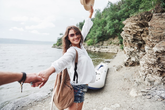 Portrait of happy young woman in hat holding man by hand and going to the boat on the lake in the mountains