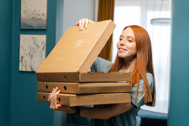 Portrait of happy young woman open boxes of hot pizza standing at doorway to apartment looking inside