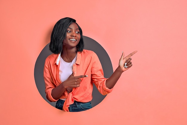 Portrait of happy young  woman smiling and pointing two fingers aside on coral background Copy space