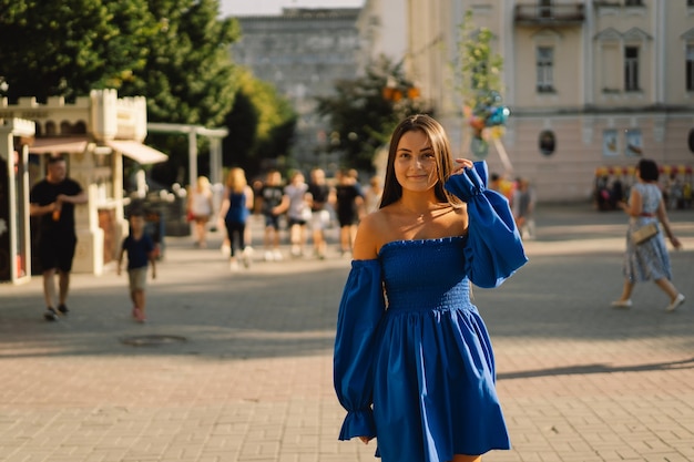 Портрет счастливой молодой женщины в синем платье, смеясь, глядя в камеру, стоя на улице городских ба ...