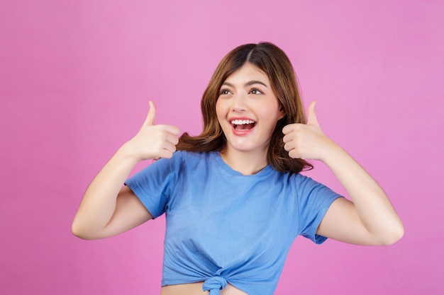 ピンクの背景の上に分離された親指を示すカジュアルなTシャツを着て幸せな若い女性の肖像画