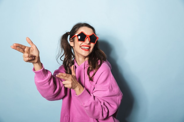 Portrait of happy young woman wearing funny sunglasses laughing and playing around over blue in studio