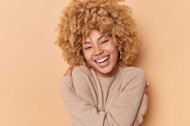 Portrait of happy young woman with curly bushy hair embraces herself feels soft dressed in casual jumper smiles broadly has white teeth isolated over beige background Self acceptance concept