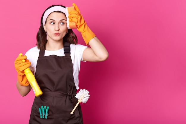 Portrait of happy young woman with orange sponge and detergent in hands, cleans her house wearing orange rubber gloves for hands protection and brown apron, isolated over pink wall. Copy space.