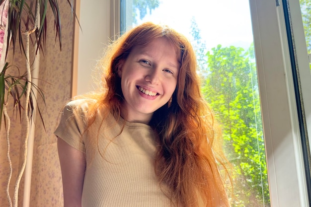 Portrait of happy young woman with red hair at home near window at sunny summer day