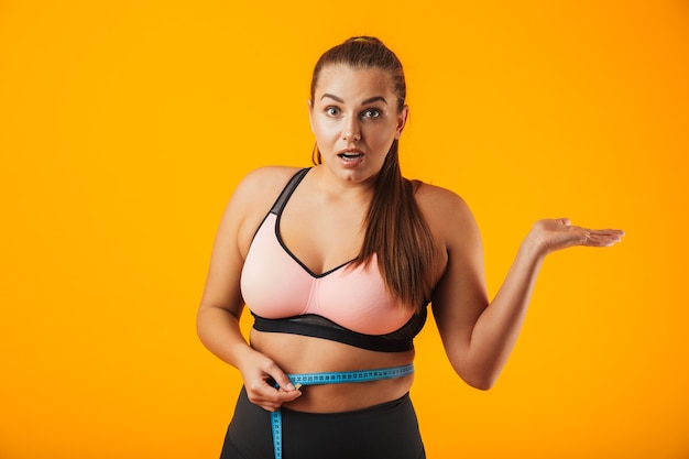 Portrait of healthy chubby woman in sportive bra measuring her waist with meter, isolated over yellow background