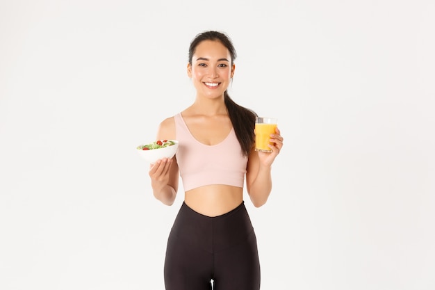 Ritratto di bella sportiva asiatica sana e in forma che tiene il succo d'arancia e insalata, colazione del mattino prima dell'allenamento, sfondo bianco.