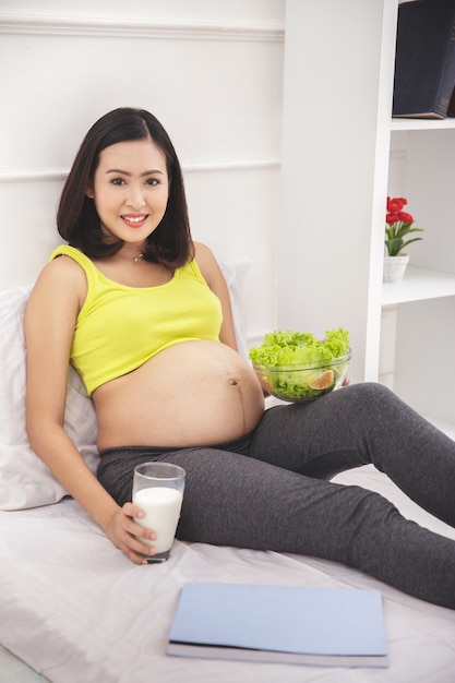 Ritratto di sana colazione donna incinta con un bicchiere di latte e una ciotola di insalata fresca in camera da letto