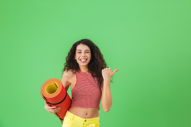 Ritratto di una donna sana 20s che sorride e porta un tappetino da yoga durante l'allenamento sulla parete verde