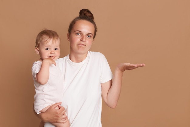 Photo portrait of helpless confused woman with dark hair wearing white t shirt standing with her baby daughter being not sure spreading hand posing isolated over brown background