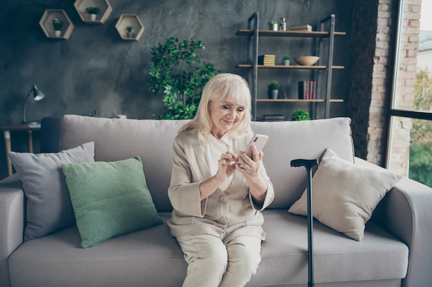 Portrait of her she nice attractive lovely cheery cheerful gray-haired grandma sitting on divan typing sms to relatives retirement at industrial brick loft modern style interior house