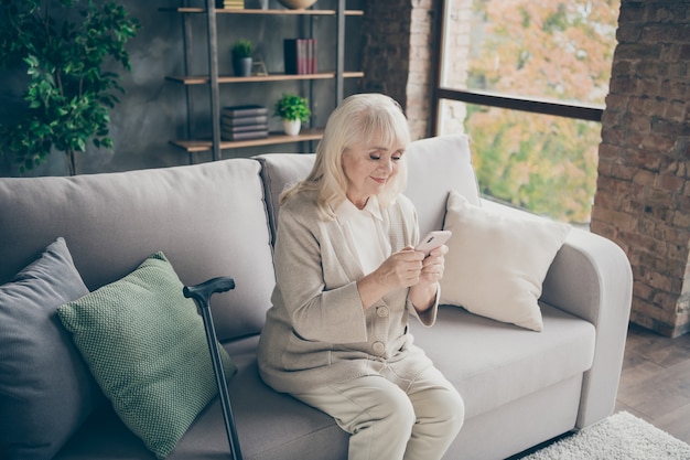 Portrait of her she nice attractive lovely focused friendly gray-haired granny sitting on divan sending sms to grandchildren spending retirement at industrial brick loft modern style interior house