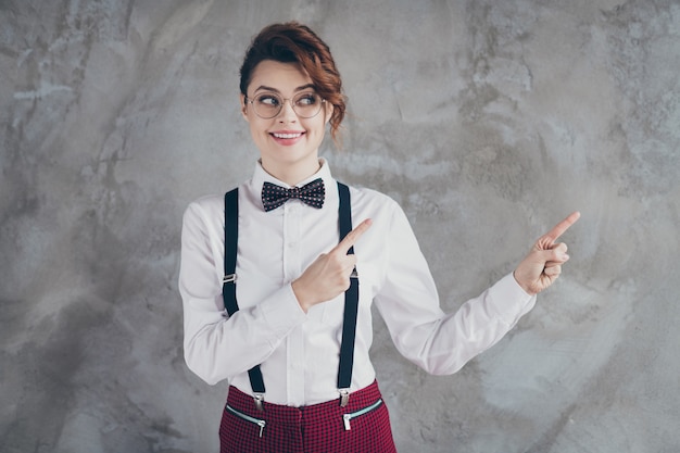 Portrait of her she nice-looking attractive lovely pretty cute\
cheerful cheery wavy-haired girl pointing two forefingers ad advert\
isolated on gray concrete industrial wall background