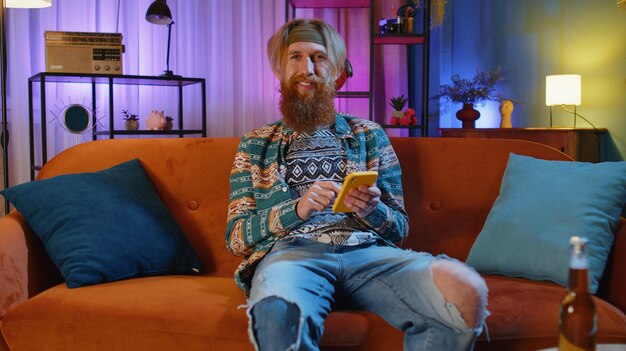 Portrait of hippie man sitting on sofa using smartphone share messages on social media application