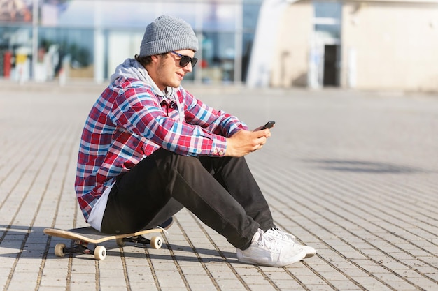 Portrait of hipster man with sunglasses holding smartphone sitting on the skateboard