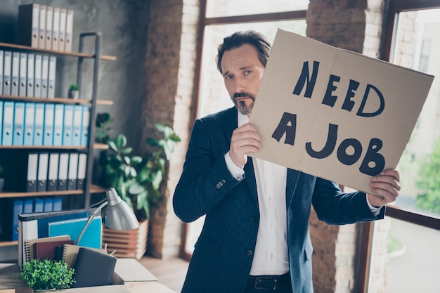 Photo portrait of his he depressed jobless guy qualified expert employer wearing jacket holding in hands placard saying need a job at modern loft industrial brick style interior workplace workstation