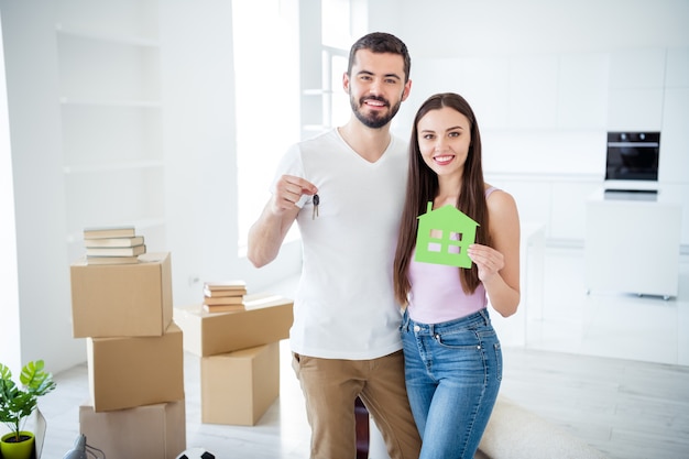 Portrait of his he her she nice attractive cheerful cheery couple embracing holding in hands key green house figure purchase trust bank loan at space flat light white interior house indoors