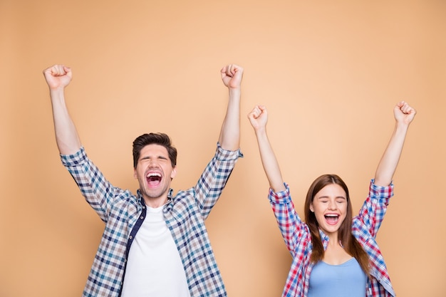 Portrait of his he her she nice attractive satisfied delighted cheerful cheery couple wearing checked shirt celebrating rising hands up good luck isolated over beige pastel color background