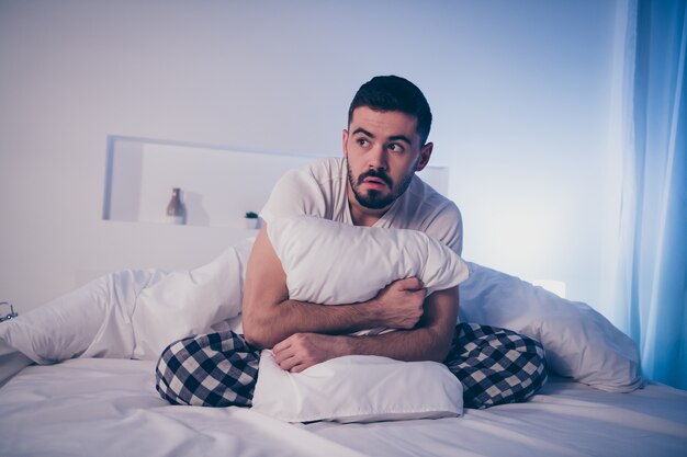 Portrait of his he nice attractive depressed scared guy sitting on bed suffering from insomnia bad time at night late evening home illuminated room flat house