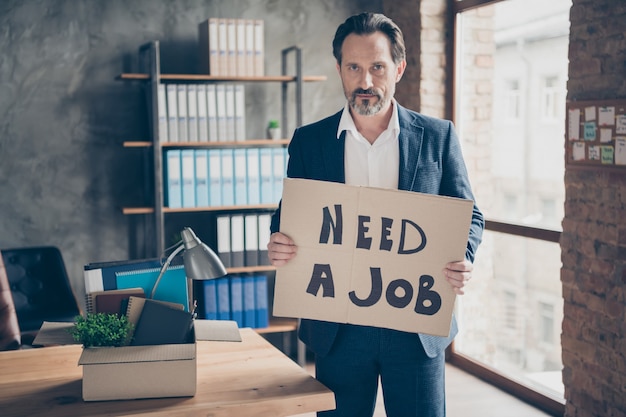 Photo portrait of his he serious jobless guy realtor economist holding in hands placard saying need a job find solution at modern loft industrial brick style interior workplace workstation