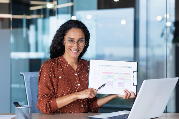 Portrait hispanic business coach woman smiling and looking at camera showing documents with economic