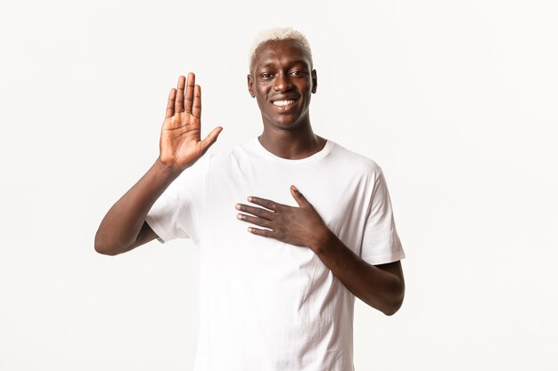 Portrait of honest attractive african-american blond guy, raising one hand and another on heart, making promise, smiling