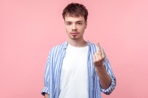 Photo portrait of hooligan, angry brown-haired man with small beard and mustache in casual striped shirt showing middle finger to haters, looking at camera. indoor studio shot isolated on pink background