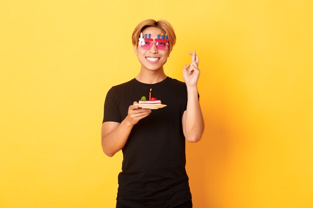 Portrait of hopeful attractive asian birthday guy, celebrating in funny party glasses, holding cake and fingers crossed while making b-day wish, yellow wall