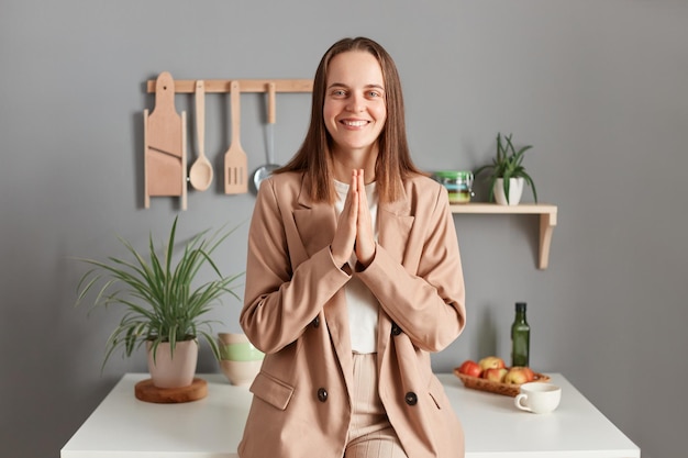 Portrait of hopeful grateful brown haired woman wearing beige suit standing near table on kitchen at home looking at camera with smile keeps palms together praying