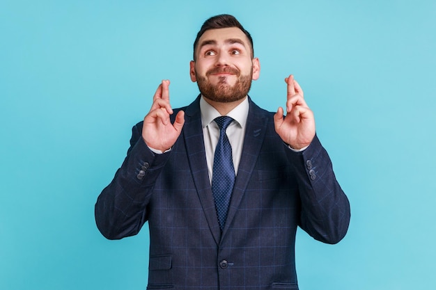 Portrait of hopeful man with beard wearing official style suit crossing fingers for luck making a wish dreaming of innermost ritual Indoor studio shot isolated on blue background
