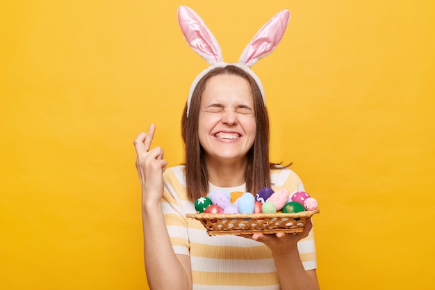 Portrait of hopeful woman wearing rabbit ears holding Easter eggs crossed fingers keeps eyes closed making wish to win Easter hunt isolated on yellow background