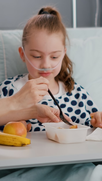 portrait of hospitalized little chid lying in bed in recovery examination room while having food meal lunch in hospital ward. sick kid eating healthy nutrition breakfest after medical surgery