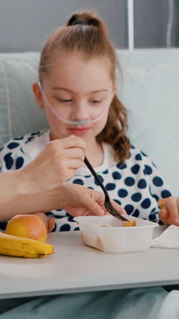 Portrait of hospitalized little chid lying in bed in recovery examination room while having food meal lunch in hospital ward. Sick kid eating healthy nutrition breakfest after medical surgery