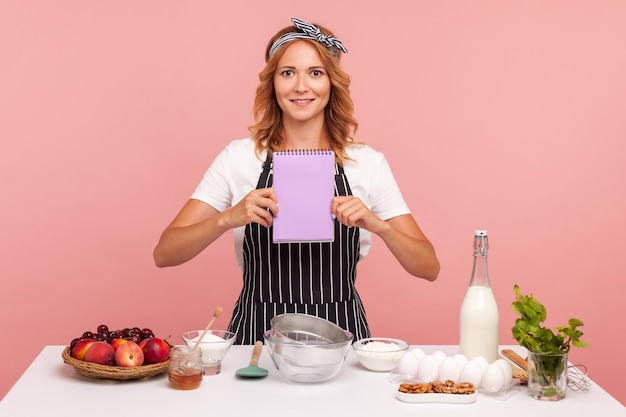 Portrait of housewife chef cook confectioner holding notebook with empty blank sheet, standing near table with bakery ingredients, copy space. Indoor studio shot isolated on pink background.