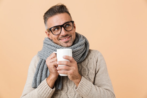 ベージュの壁に隔離された熱いお茶のカップを保持しているセーターとスカーフに身を包んだ幸せな男が目をそらしている場合の肖像画