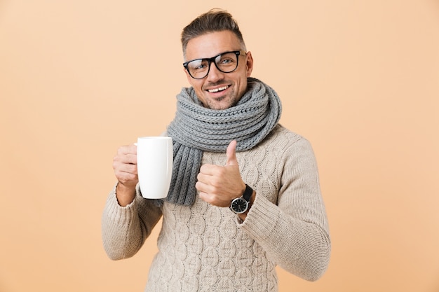 Portrait if a happy man dressed in sweater and scarf holding cup of hot tea isolated over beige wall