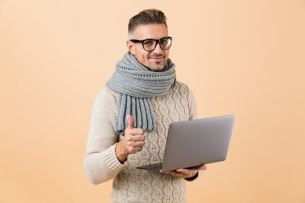 Ritratto se un uomo felice vestito in maglione e sciarpa in piedi isolato sul muro beige, tenendo il computer portatile, dando i pollici in su