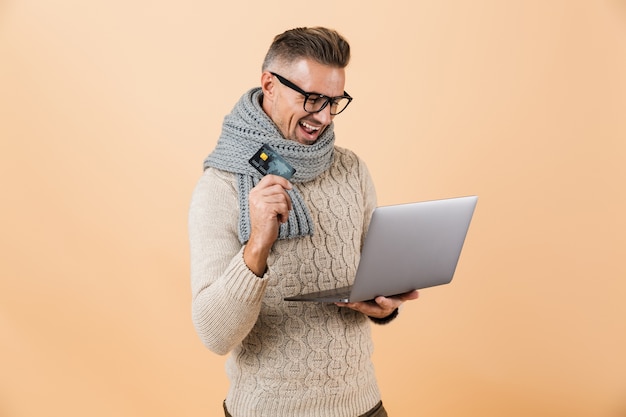 Ritratto se un uomo felice vestito in maglione e sciarpa in piedi isolato sul muro beige, tenendo il computer portatile, mostrando la carta di credito in plastica