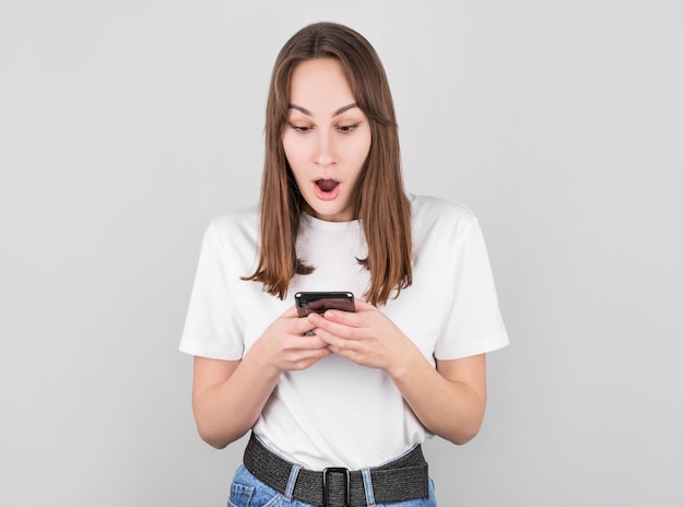 Portrait if a shocked young girl looking at mobile phone isolated over gray wall