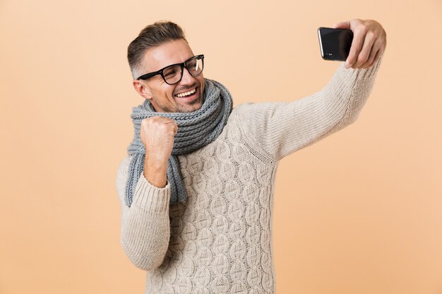 Portrait if a smiling man dressed in sweater and scarf standing isolated over beige wall, taking a selfie, celebrating