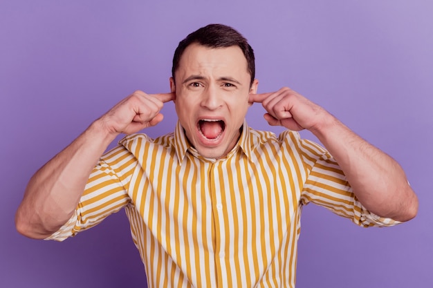 Portrait of ignoring crazy mad guy fingers close ears yell open mouth on violet background