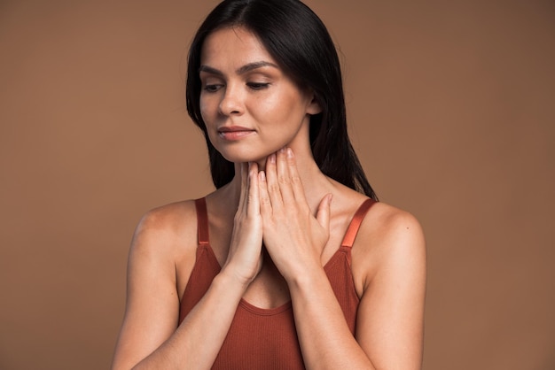 Portrait of ill woman with brown hair standing and touching her neck while frowning from pain, suffering sore throat, flu symptom. Indoor studio shot isolated on brown background