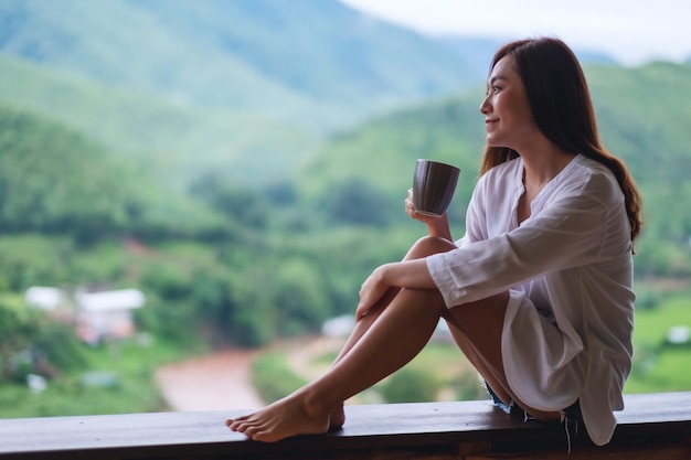 山と緑の自然を見ながら、バルコニーでホットコーヒーを保持し、飲む美しいアジアの女性の肖像画の画像