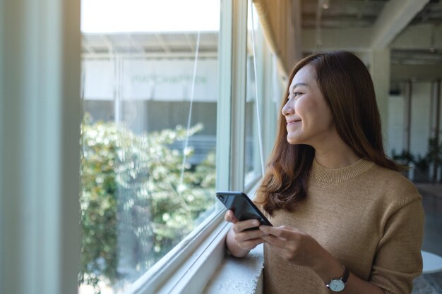 携帯電話を持って使用しながら窓の外を見ている美しい若いアジアの女性の肖像画の画像