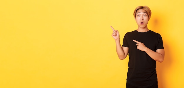 Portrait of impressed and excited handsome asian guy in black tshirt react to your logo pointing fin...