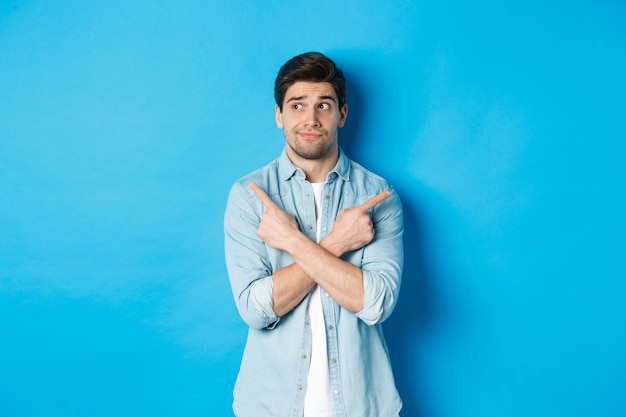 Portrait of indecisive adult man pointing fingers sideways but looking left, making choice between two products, standing against blue wall