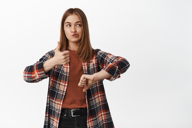 Foto ritratto di ragazza indecisa mostra antipatia o simile, pollice in alto e pollice in giù, guardando da parte pensieroso, pensando alla valutazione, giudicando smth, in piedi su sfondo bianco