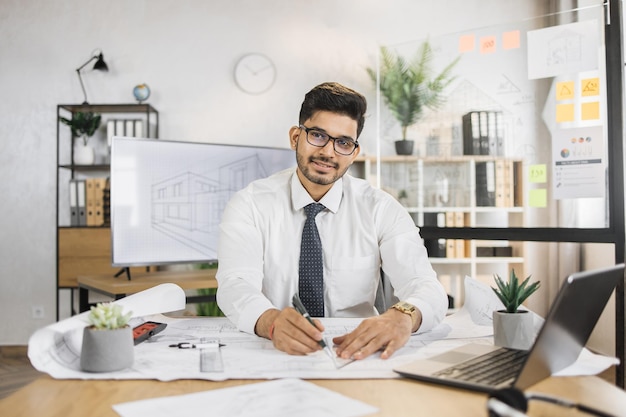 Portrait of indian man wearing formal wear and hard hat at work in modern office