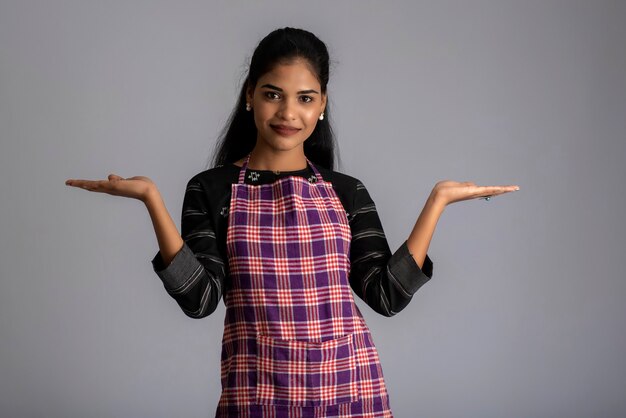 Portrait of Indian woman chef or cook in an apron, presenting, pointing, with ok sign, thumbs up on the grey.