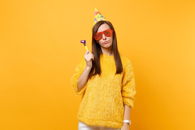 Portrait of indifferent woman in orange funny glasses, birthday hat with playing pipe standing celebrating isolated on yellow background. People sincere emotions, lifestyle concept. Advertising area.